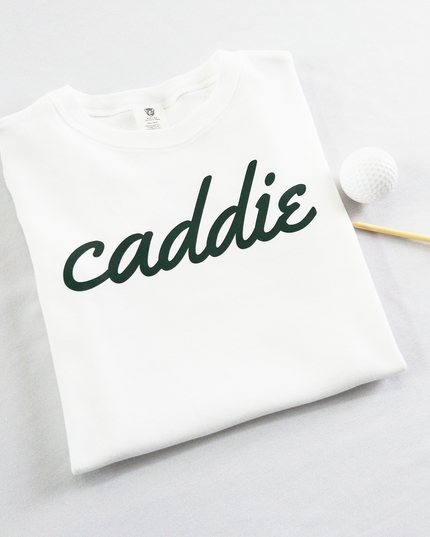 White kids golf t-shirt with dark green cursive text reading 'caddie,' displayed flat with golf balls and a wooden tee, perfect for young golf enthusiasts and future caddies.