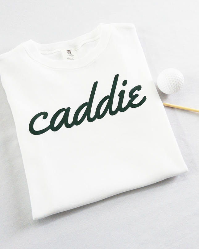 White kids golf t-shirt with dark green cursive text reading 'caddie,' displayed flat with golf balls and a wooden tee, perfect for young golf enthusiasts and future caddies.