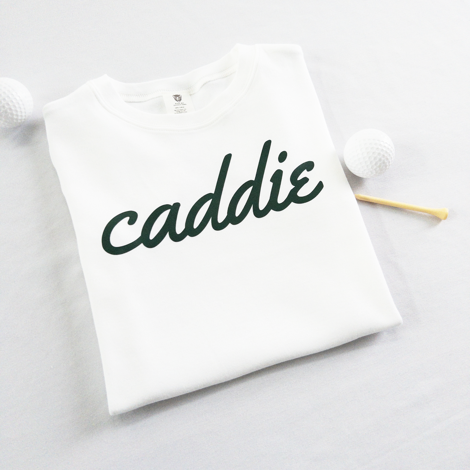 White kids golf t-shirt with dark green cursive text reading 'caddie,' displayed flat with golf balls and a wooden tee, perfect for young golf enthusiasts and future caddies.