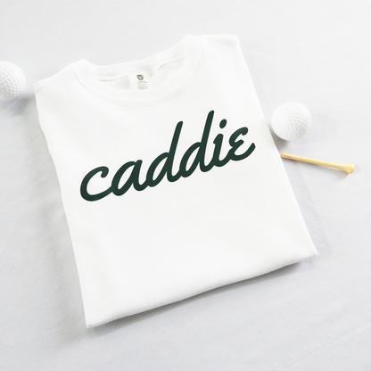 White kids golf t-shirt with dark green cursive text reading 'caddie,' displayed flat with golf balls and a wooden tee, perfect for young golf enthusiasts and future caddies.