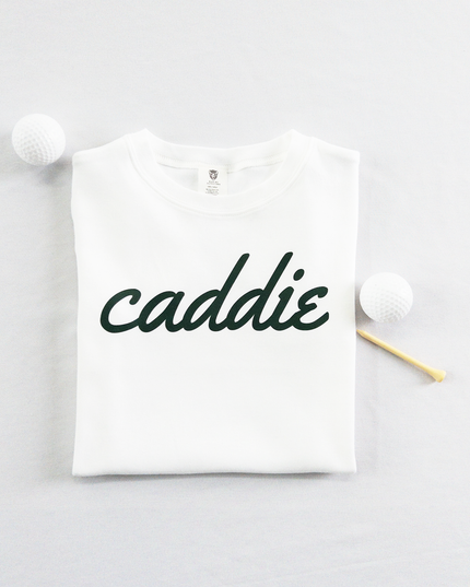 White kids golf t-shirt with dark green cursive text reading 'caddie,' displayed flat with golf balls and a wooden tee, perfect for young golf enthusiasts and future caddies.