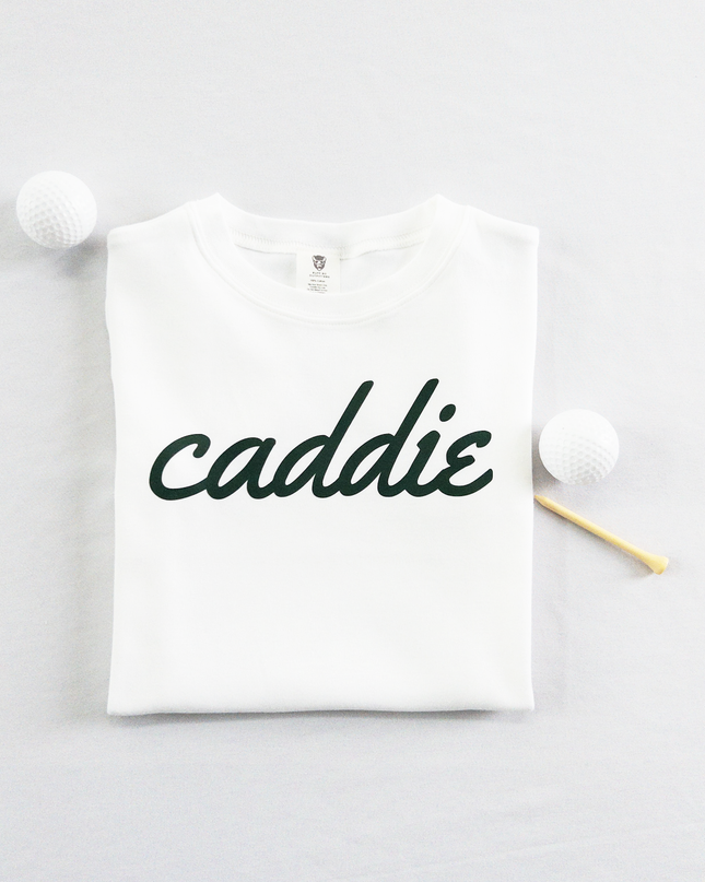 White kids golf t-shirt with dark green cursive text reading 'caddie,' displayed flat with golf balls and a wooden tee, perfect for young golf enthusiasts and future caddies.