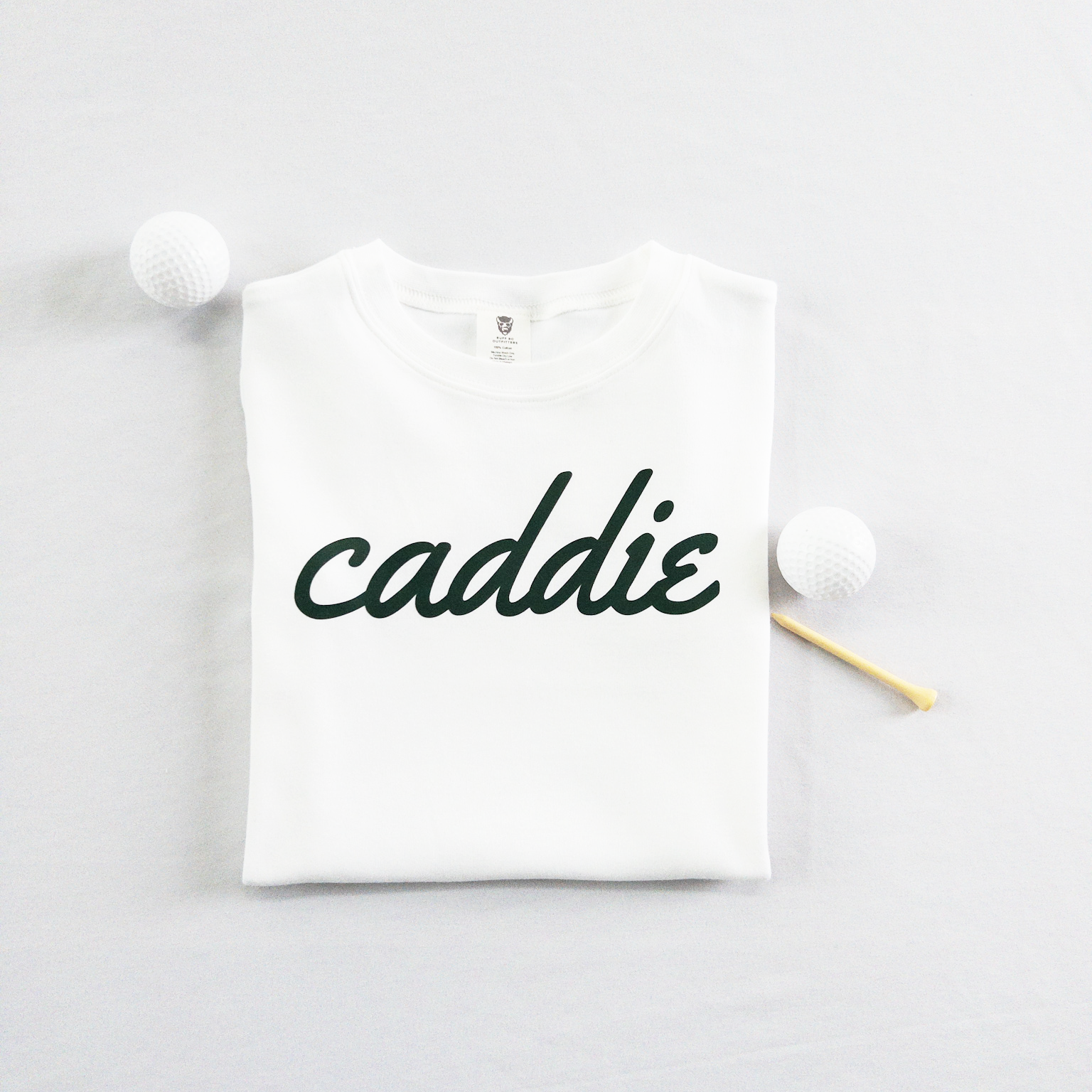 White kids golf t-shirt with dark green cursive text reading 'caddie,' displayed flat with golf balls and a wooden tee, perfect for young golf enthusiasts and future caddies.