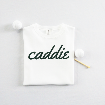 White kids golf t-shirt with dark green cursive text reading 'caddie,' displayed flat with golf balls and a wooden tee, perfect for young golf enthusiasts and future caddies.