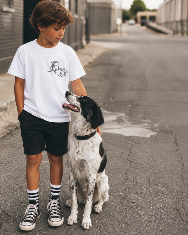 A young boy wearing a white graphic t-shirt, black shorts, and striped socks stands on an urban street next to a black and white dog.