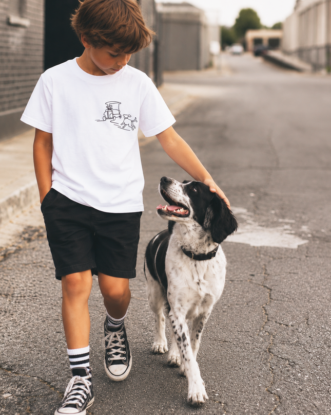 A boy walking a black and white dog down an urban street, wearing a white t-shirt with a small black graphic of a dog wearing a top hat, black shorts, and black and white sneakers.