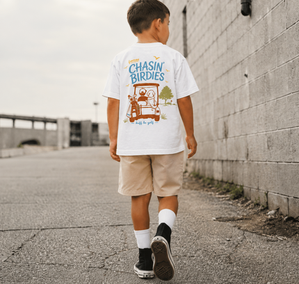 A boy seen from behind walking down an outdoor alleyway, wearing a white graphic t-shirt with a cartoon birdie design and the text 'CHASIN' BIRDIES', paired with beige shorts, white socks, and black sneakers.