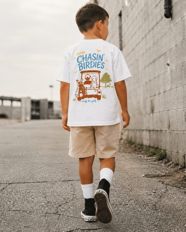 A boy seen from behind walking down an outdoor alleyway, wearing a white graphic t-shirt with a cartoon birdie design and the text 'CHASIN' BIRDIES', paired with beige shorts, white socks, and black sneakers.