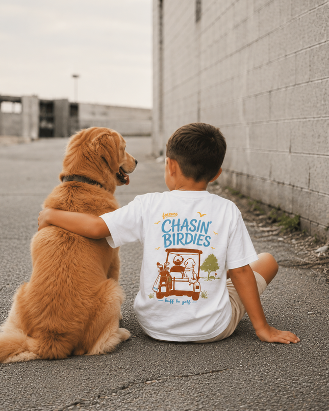 A young boy and his golden retriever sit together on a paved street next to a gray brick wall, viewed from behind. The boy is wearing a white graphic t-shirt with a colorful illustration and the text 'Chasin' Birdies' on the back.