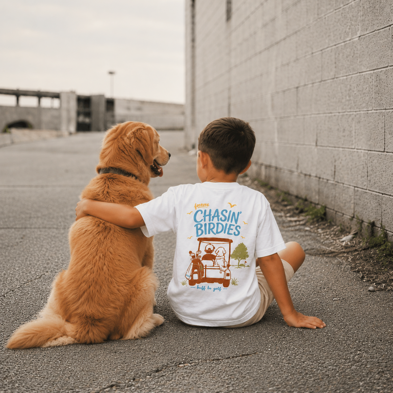 A young boy and his golden retriever sit together on a paved street next to a gray brick wall, viewed from behind. The boy is wearing a white graphic t-shirt with a colorful illustration and the text 'Chasin' Birdies' on the back.