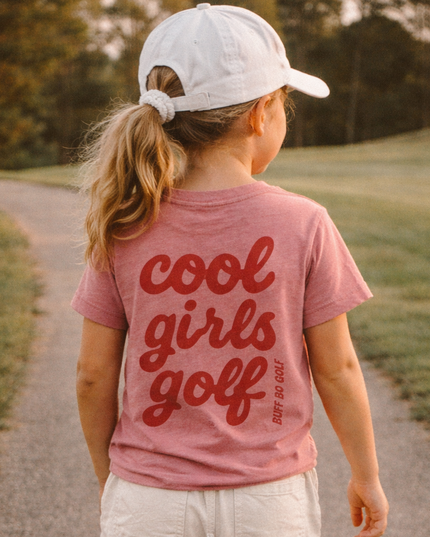 Child wearing a 'cool girls golf' t-shirt walking on a path outdoors.