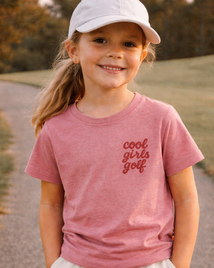 Young girl wearing a pink 'cool girls golf' t-shirt and white cap standing on a path outdoors.