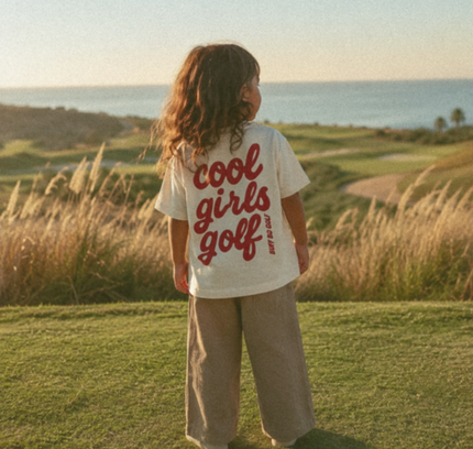 Child wearing a 'cool girls golf' shirt standing on a golf course with ocean view