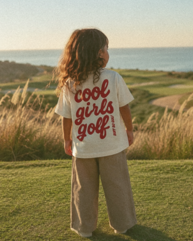 Child wearing a 'cool girls golf' shirt standing on a golf course with ocean view