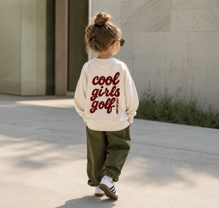 A child seen from behind wearing a cream sweatshirt with 'cool girls golf' printed in dark red script on the back, paired with olive green pants and white sneakers, walking outdoors on a sunny day.