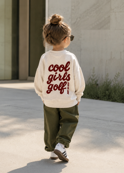 A child seen from behind wearing a cream sweatshirt with 'cool girls golf' printed in dark red script on the back, paired with olive green pants and white sneakers, walking outdoors on a sunny day.