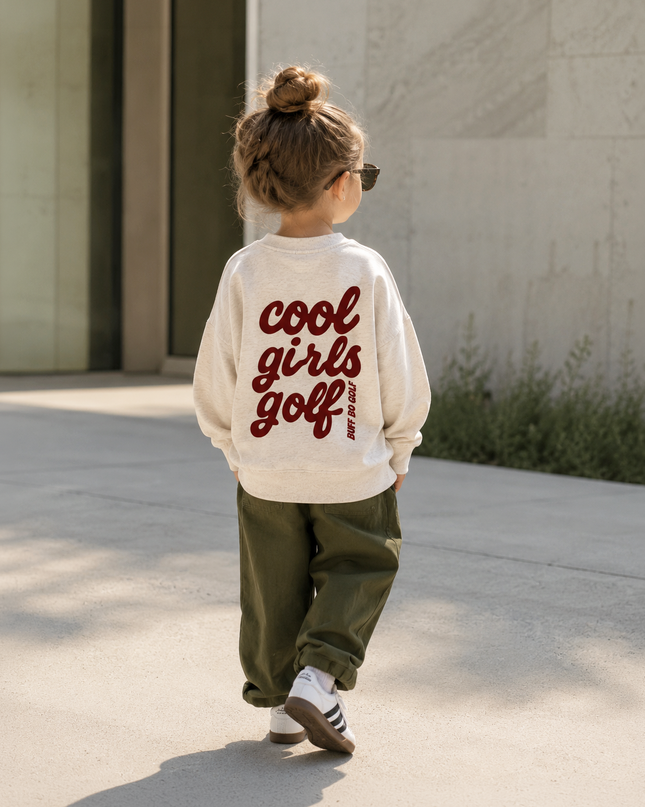 A child seen from behind wearing a cream sweatshirt with 'cool girls golf' printed in dark red script on the back, paired with olive green pants and white sneakers, walking outdoors on a sunny day.