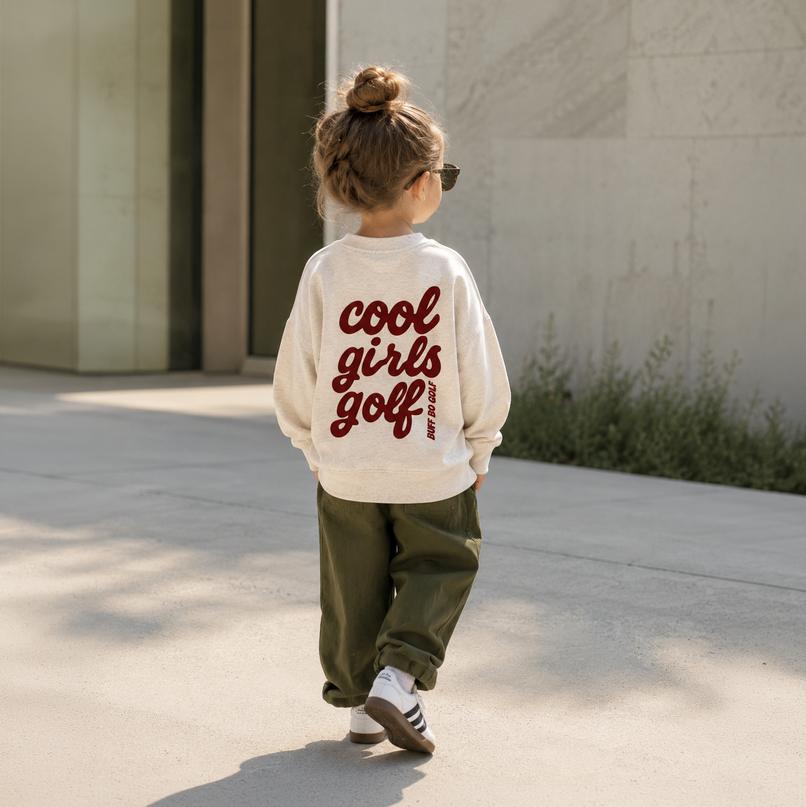 A child seen from behind wearing a cream sweatshirt with 'cool girls golf' printed in dark red script on the back, paired with olive green pants and white sneakers, walking outdoors on a sunny day.