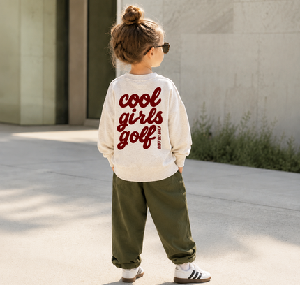 A child standing outdoors on a sunny day, viewed from behind, wearing a cream long-sleeve sweatshirt with 'cool girls golf' printed in red across the back, olive green pants, and white sneakers.