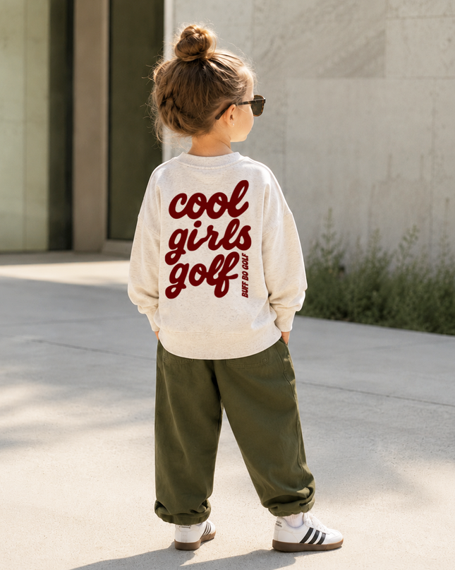 A child standing outdoors on a sunny day, viewed from behind, wearing a cream long-sleeve sweatshirt with 'cool girls golf' printed in red across the back, olive green pants, and white sneakers.