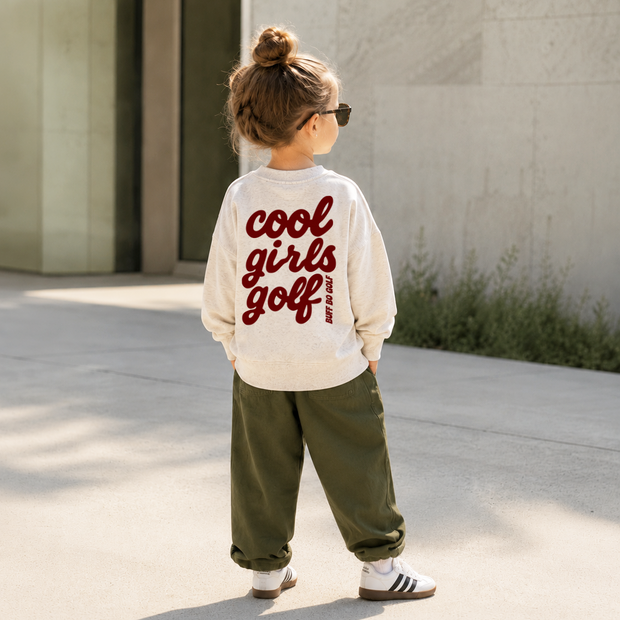 A child standing outdoors on a sunny day, viewed from behind, wearing a cream long-sleeve sweatshirt with 'cool girls golf' printed in red across the back, olive green pants, and white sneakers.