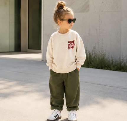 A young child wearing a cream sweatshirt with red text that reads 'cool girl golf', olive green pants, and white sneakers, standing outdoors in front of a modern building.
