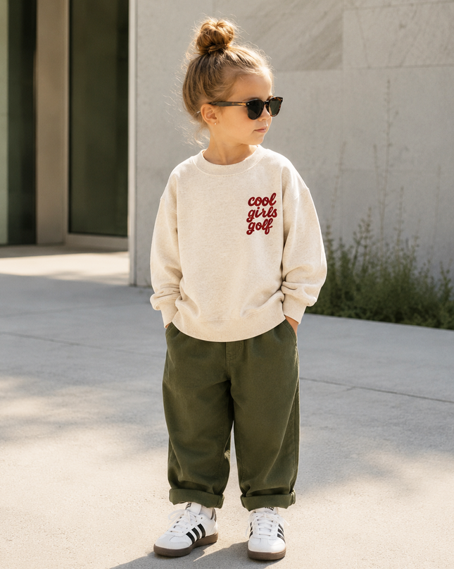 A young child wearing a cream sweatshirt with red text that reads 'cool girl golf', olive green pants, and white sneakers, standing outdoors in front of a modern building.