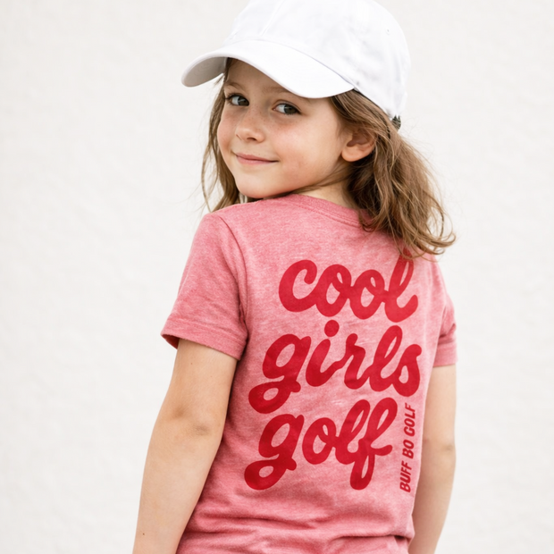 Young girl wearing a pink 'cool girls golf' t-shirt and white cap on a plain background