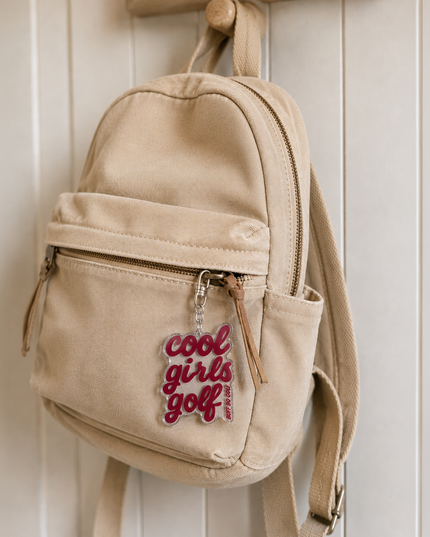 A beige canvas mini backpack hanging on a wooden peg against a white paneled wall. The backpack features a front zippered pocket with a silver zipper pull that reads 'cool girls golf' in red script.
