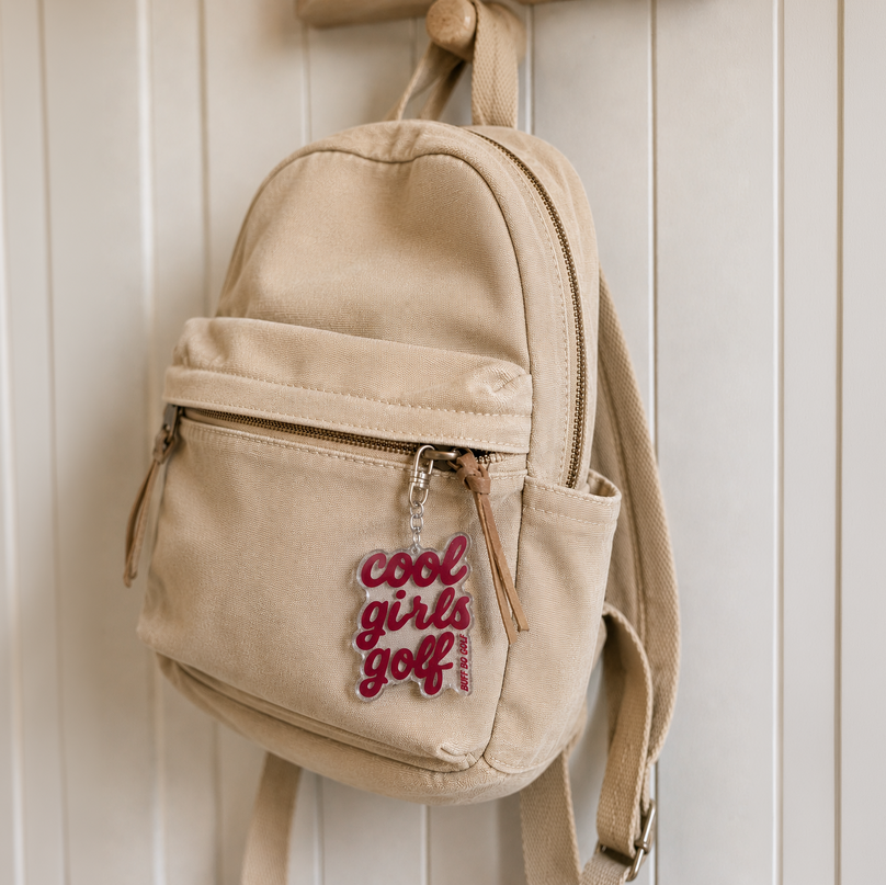 A beige canvas mini backpack hanging on a wooden peg against a white paneled wall. The backpack features a front zippered pocket with a silver zipper pull that reads 'cool girls golf' in red script.
