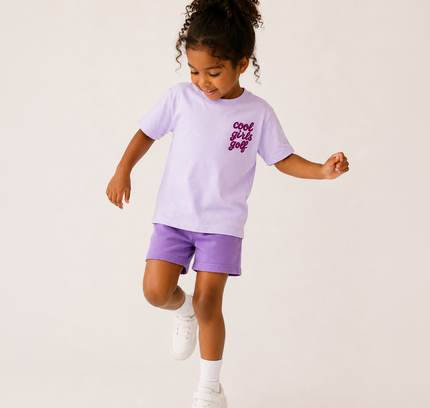 A young girl with curly hair is captured mid-dance in a studio setting, wearing a light purple t-shirt with 'cool girls just' printed in dark purple on the chest, matching purple shorts, white socks, and white sneakers against a solid off-white background.
