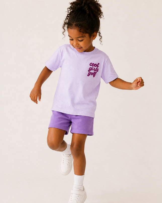 A young girl with curly hair is captured mid-dance in a studio setting, wearing a light purple t-shirt with 'cool girls just' printed in dark purple on the chest, matching purple shorts, white socks, and white sneakers against a solid off-white background.