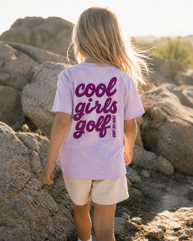 A young girl seen from behind wearing a light lavender t-shirt with 'cool girls golf' printed in dark purple across the back, standing outdoors among large rocks in a sunny, rocky desert landscape.