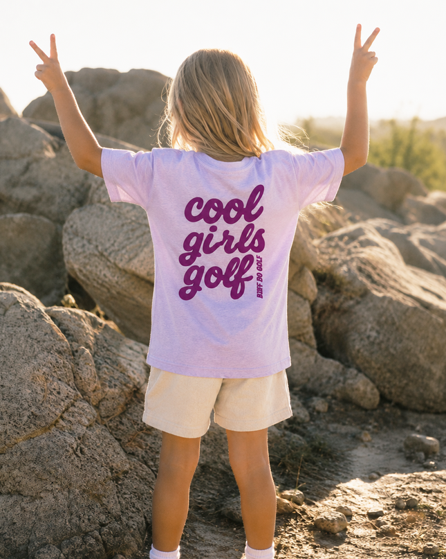 A young girl stands with her back to the camera, posing with peace signs in front of large rocks outdoors. She is wearing a light lavender t-shirt with 'cool girls golf' printed in dark purple across the back, paired with cream shorts.