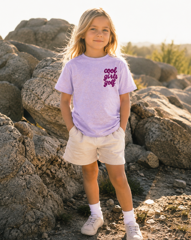 A young girl stands outdoors among large rocks in warm golden-hour light, wearing a light lavender graphic T-shirt with purple text reading 'cool girls go' and cream shorts.