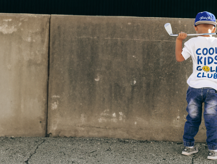 Child holding golf clubs against a concrete wall wearing a 'Cool Kids Golf Club' shirt.