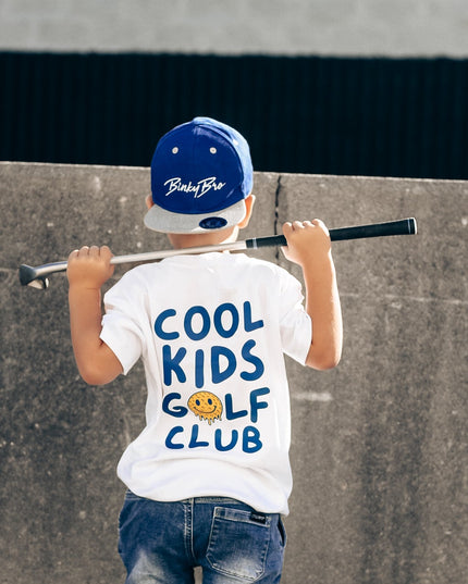 Child wearing a 'Cool Kids Golf Club' t-shirt and blue cap, standing against a concrete wall.
