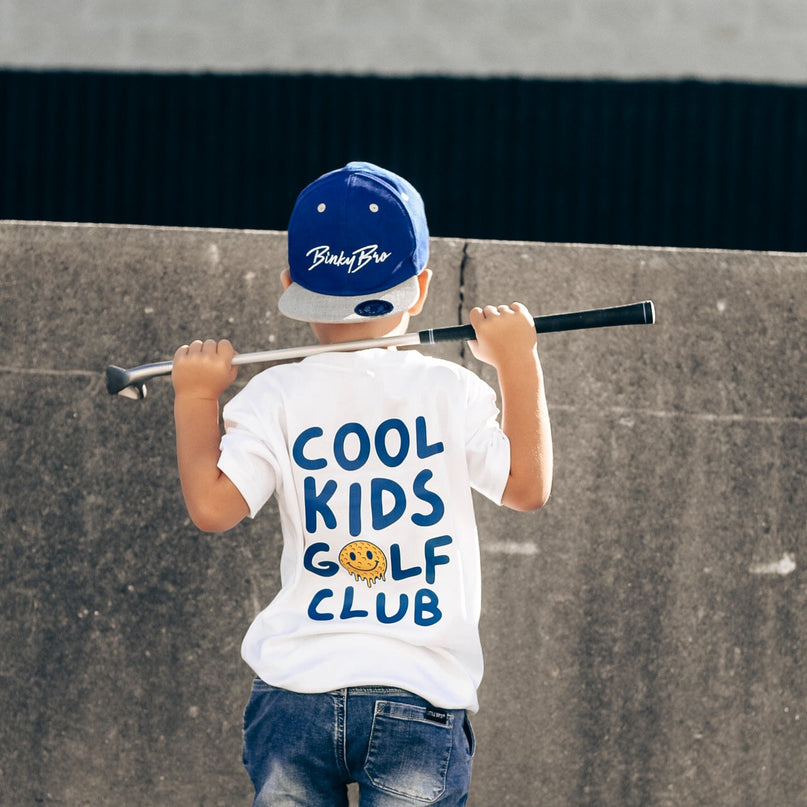 Child wearing a 'Cool Kids Golf Club' t-shirt and blue cap, standing against a concrete wall.