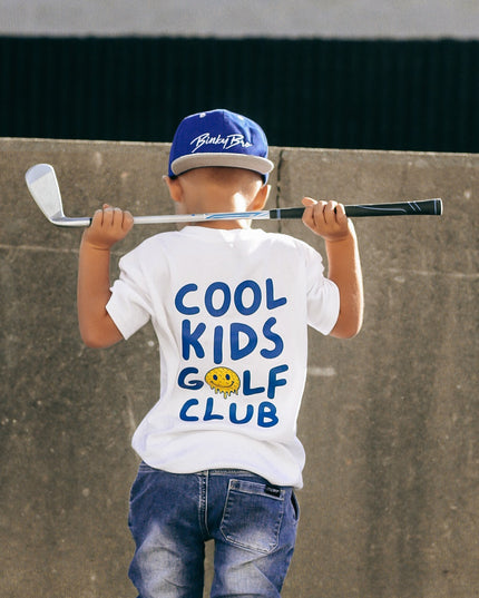 Child wearing a 'Cool Kids Golf Club' t-shirt holding a golf club against a concrete wall.