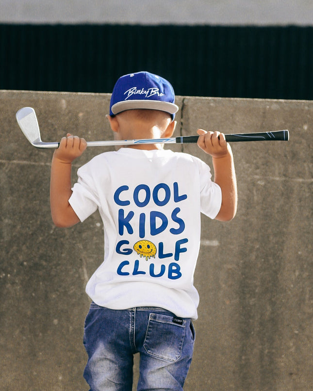 Child wearing a 'Cool Kids Golf Club' t-shirt holding a golf club against a concrete wall.