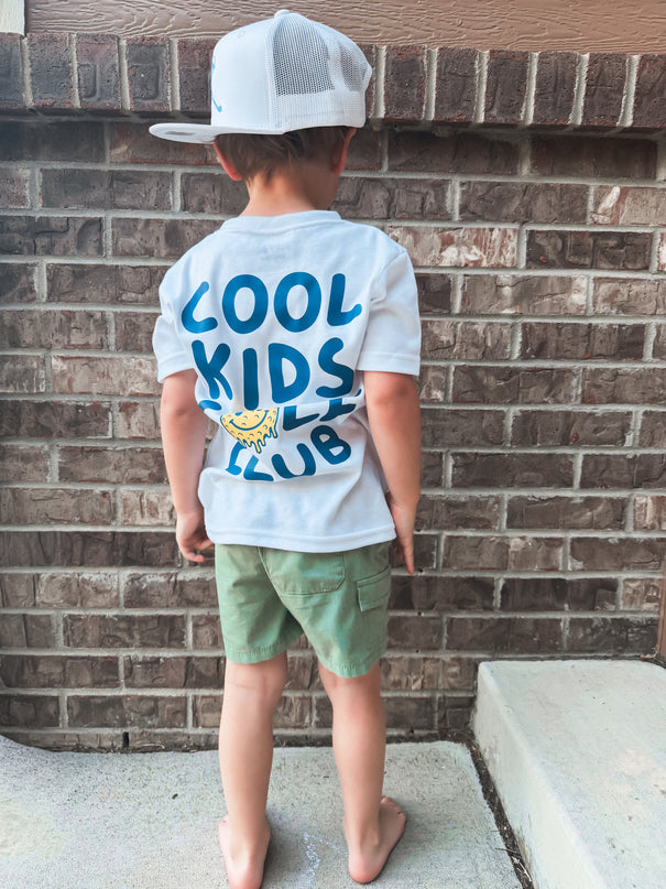 Child wearing a 'Cool Kids Club' t-shirt standing against a brick wall.