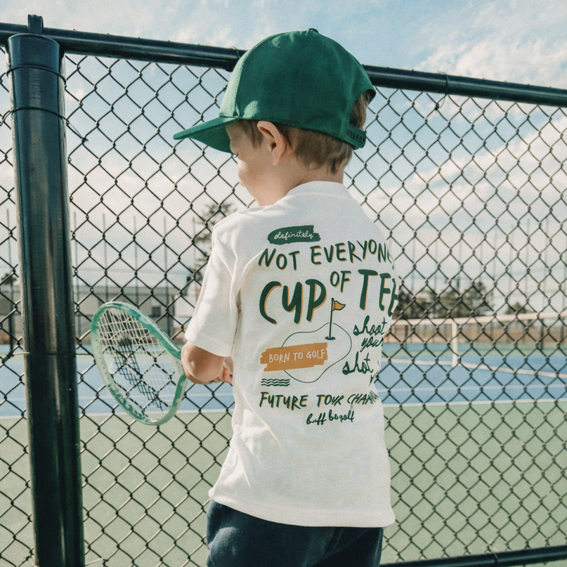 Child wearing a white t-shirt with text and a green cap on a tennis court.