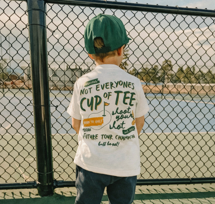 Child wearing a white t-shirt with text, standing near a chain-link fence with a scenic background.