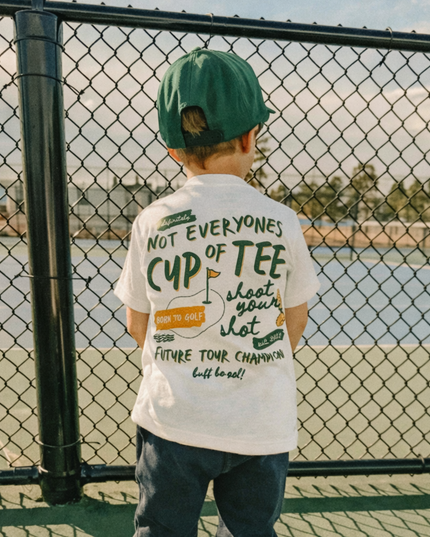 Child wearing a white t-shirt with text, standing near a chain-link fence with a scenic background.