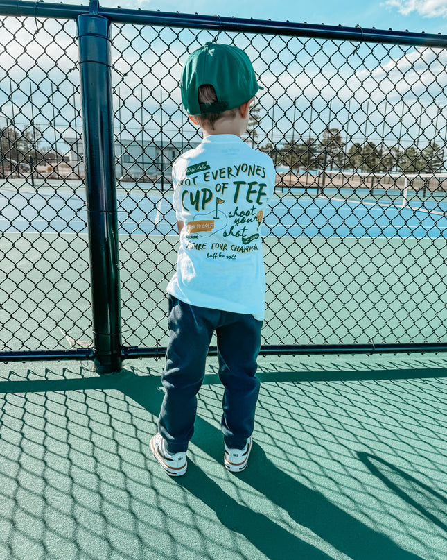 Child wearing a light blue t-shirt with text, standing on a tennis court.