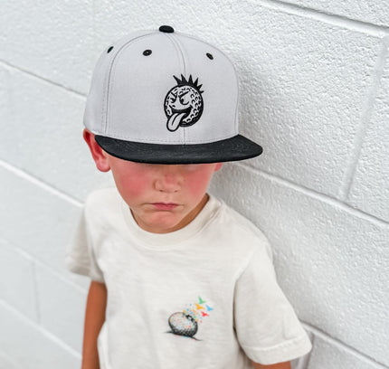 Child wearing a cap with a logo and a t-shirt with a small design, standing against a white wall.