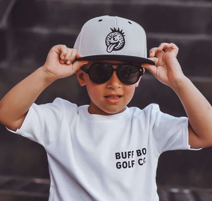 Child wearing a white t-shirt with text, sunglasses, and a cap on a dark background