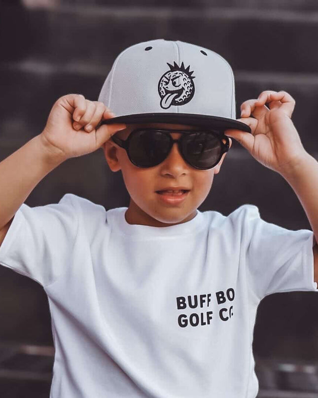 Child wearing a white t-shirt with text, sunglasses, and a cap on a dark background