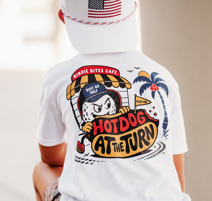 A child wearing a white graphic t-shirt, camouflage shorts, and a white cap with an American flag patch, shown from behind. The t-shirt features a large cartoon hot dog illustration with the text 'Hirpie Bites Cafe' and 'Hotdog at the Turn'.
