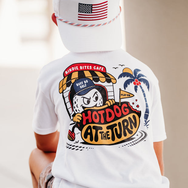 A child wearing a white graphic t-shirt, camouflage shorts, and a white cap with an American flag patch, shown from behind. The t-shirt features a large cartoon hot dog illustration with the text 'Hirpie Bites Cafe' and 'Hotdog at the Turn'.